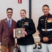 Three individuals pose for a formal photograph at what appears to be a military awards ceremony. A female Marine in dress blues holds a plaque award, flanked by a civilian in a brown suit and a senior enlisted Marine in dress blues with extensive medals and chevrons. The plaque appears to be from the United States Marine Corps Correspondents Association for the year 2023.