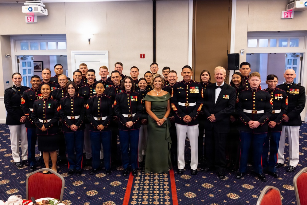 A formal group photograph of approximately 30 United States Marines in dress blue uniforms, posing with two civilians — a woman in an olive green evening gown and a man in a tuxedo — in what appears to be a banquet or event hall. The setting and attire suggest this is a formal military ceremony or ball, likely a Marine Corps Birthday Ball or similar commemorative event. The group is arranged in multiple rows against a neutral-colored wall with EXIT signs and windows visible in the background.