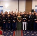 A formal group photograph of approximately 30 United States Marines in dress blue uniforms, posing with two civilians — a woman in an olive green evening gown and a man in a tuxedo — in what appears to be a banquet or event hall. The setting and attire suggest this is a formal military ceremony or ball, likely a Marine Corps Birthday Ball or similar commemorative event. The group is arranged in multiple rows against a neutral-colored wall with EXIT signs and windows visible in the background.
