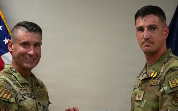 Two U.S. Air Force personnel in OCP (Operational Camouflage Pattern) uniforms are posing together while jointly holding a Department of the Air Force certificate. The ceremony appears to be an award presentation, as the service member on the right is wearing a medal with a blue and gold ribbon. The setting includes an American flag and what appears to be a Maine Air National Guard flag in the background.