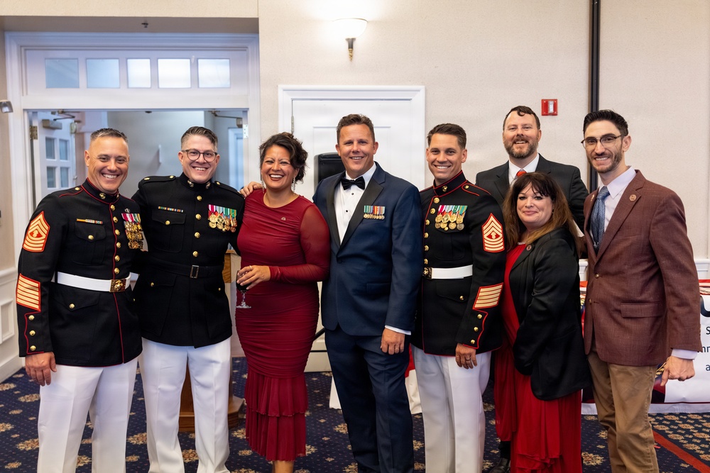 A group of eight people posing together at what appears to be a formal military event or gala. Three individuals are wearing United States Marine Corps dress blue uniforms with medals and white trousers, while the others are dressed in formal civilian attire including suits and evening wear. The setting appears to be a banquet hall or event venue.