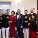 A group of eight people posing together at what appears to be a formal military event or gala. Three individuals are wearing United States Marine Corps dress blue uniforms with medals and white trousers, while the others are dressed in formal civilian attire including suits and evening wear. The setting appears to be a banquet hall or event venue.