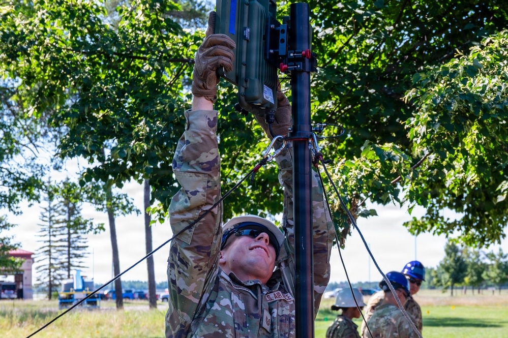 A U.S. Air Force service member in OCP (Operational Camouflage Pattern) uniform is erecting or adjusting a communications or antenna mast in an outdoor field environment. The soldier is reaching upward with both gloved hands to secure equipment at the top of a telescoping pole stabilized by guy wires. Additional personnel in blue hard hats are visible in the background.