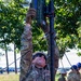A U.S. Air Force service member in OCP (Operational Camouflage Pattern) uniform is erecting or adjusting a communications or antenna mast in an outdoor field environment. The soldier is reaching upward with both gloved hands to secure equipment at the top of a telescoping pole stabilized by guy wires. Additional personnel in blue hard hats are visible in the background.