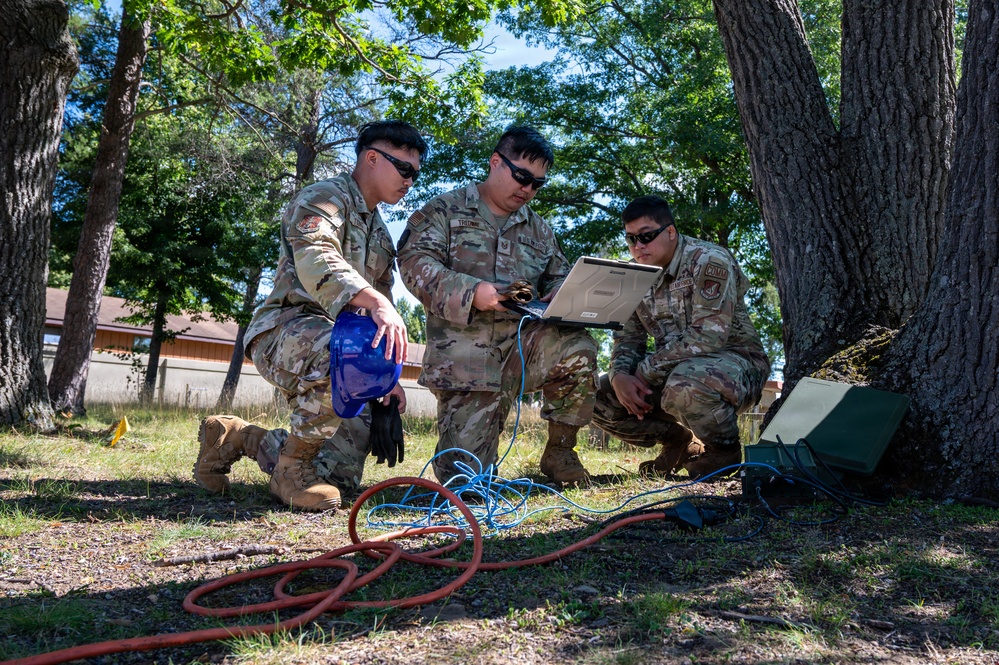 Three military personnel in OCP (Operational Camouflage Pattern) uniforms are gathered outdoors under trees, examining a ruggedized laptop connected to multiple cables. They appear to be conducting a field communications or technical operations exercise, with various colored cables (red, blue, orange) running along the ground connected to a green military equipment case. The setting appears to be a training area or base with trees and a building visible in the background.