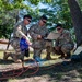 Three military personnel in OCP (Operational Camouflage Pattern) uniforms are gathered outdoors under trees, examining a ruggedized laptop connected to multiple cables. They appear to be conducting a field communications or technical operations exercise, with various colored cables (red, blue, orange) running along the ground connected to a green military equipment case. The setting appears to be a training area or base with trees and a building visible in the background.