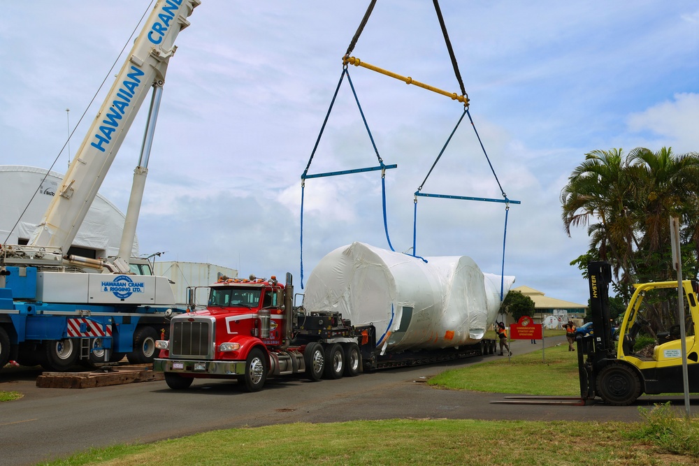 A heavy-lift crane operation is underway at what appears to be an industrial or port facility in Hawaii. A large, shrink-wrapped cylindrical object is being lifted by a 'Hawaiian Crane &amp; Rigging Ltd.' crane from a red semi-truck flatbed trailer. A yellow Hyster forklift and several workers are visible on site assisting with the operation.