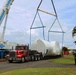 A heavy-lift crane operation is underway at what appears to be an industrial or port facility in Hawaii. A large, shrink-wrapped cylindrical object is being lifted by a 'Hawaiian Crane &amp; Rigging Ltd.' crane from a red semi-truck flatbed trailer. A yellow Hyster forklift and several workers are visible on site assisting with the operation.