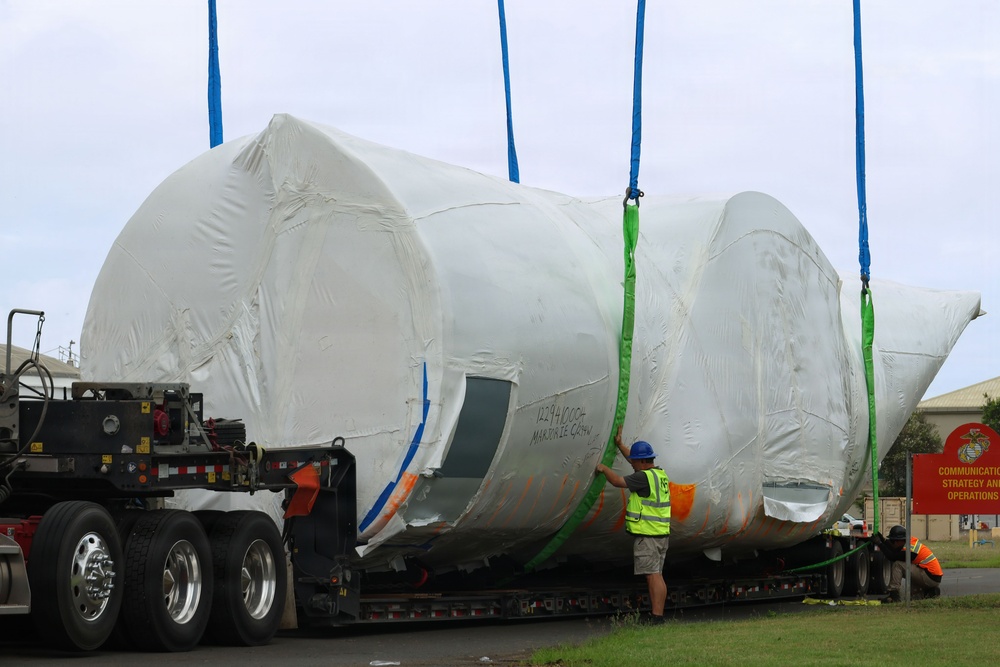A large aircraft fuselage or aerospace component, wrapped in white protective shrink wrap, is being transported on a flatbed semi-trailer. Workers in high-visibility safety vests and hard hats are managing lifting straps (blue and green) attached to the wrapped object. The scene appears to be at or near a U.S. Marine Corps facility, as indicated by a red sign visible in the background.