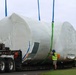 A large aircraft fuselage or aerospace component, wrapped in white protective shrink wrap, is being transported on a flatbed semi-trailer. Workers in high-visibility safety vests and hard hats are managing lifting straps (blue and green) attached to the wrapped object. The scene appears to be at or near a U.S. Marine Corps facility, as indicated by a red sign visible in the background.