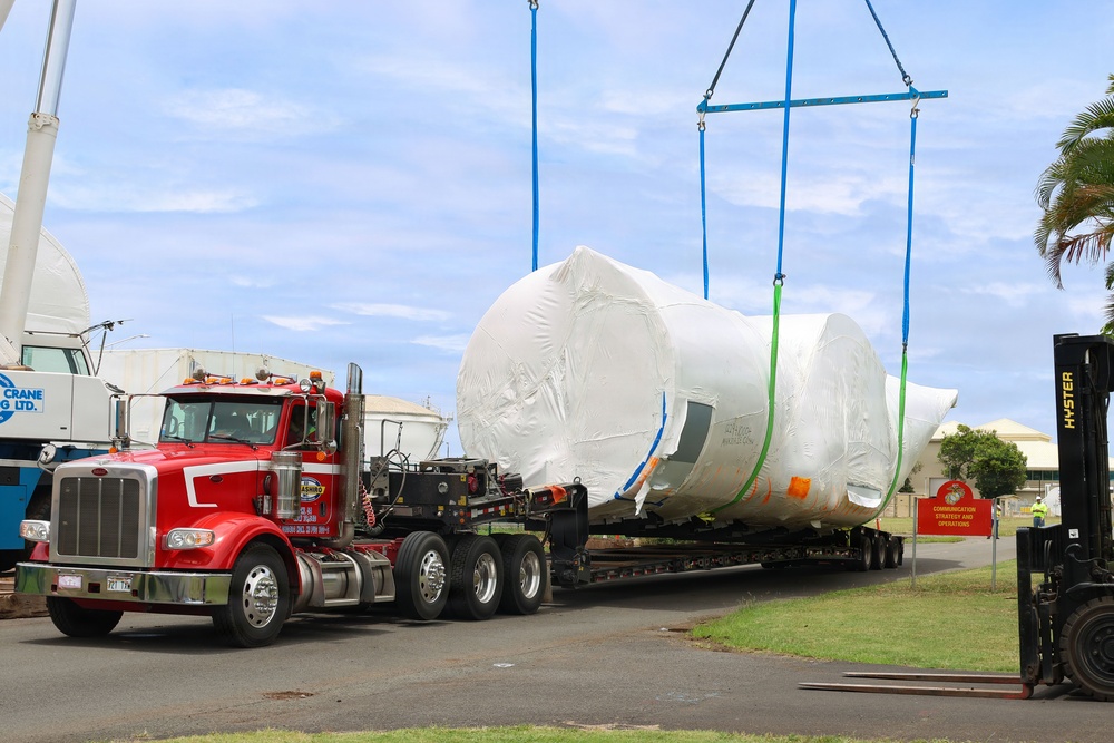 A large oversized load wrapped in white protective plastic sheeting is being lifted by crane straps from a red semi-truck flatbed trailer. The operation appears to be taking place at an industrial or military facility, with a Hyster forklift visible on the right and crane equipment on the left. The wrapped cargo appears to be a large cylindrical or vessel-shaped object, possibly industrial equipment or a large tank.