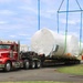 A large oversized load wrapped in white protective plastic sheeting is being lifted by crane straps from a red semi-truck flatbed trailer. The operation appears to be taking place at an industrial or military facility, with a Hyster forklift visible on the right and crane equipment on the left. The wrapped cargo appears to be a large cylindrical or vessel-shaped object, possibly industrial equipment or a large tank.