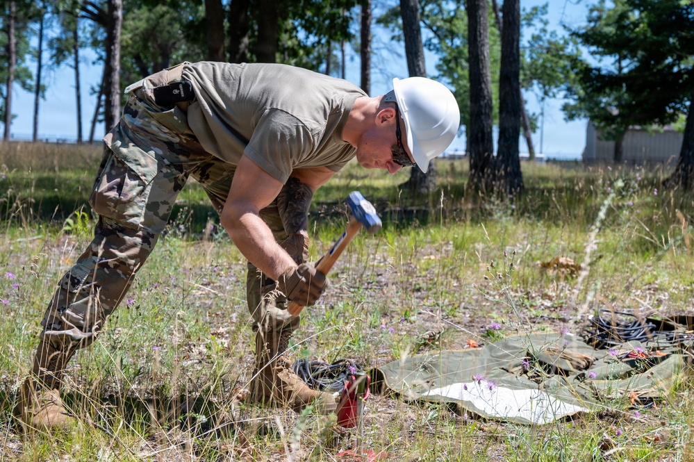 A military service member wearing camouflage uniform and a white hard hat is using a hammer or mallet to drive something into the ground in an outdoor, grassy area. The individual is bent over in a striking posture, working near what appears to be equipment laid out on a tarp or ground cloth. The setting appears to be a wooded training or work area during daytime.