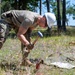 A military service member wearing camouflage uniform and a white hard hat is using a hammer or mallet to drive something into the ground in an outdoor, grassy area. The individual is bent over in a striking posture, working near what appears to be equipment laid out on a tarp or ground cloth. The setting appears to be a wooded training or work area during daytime.