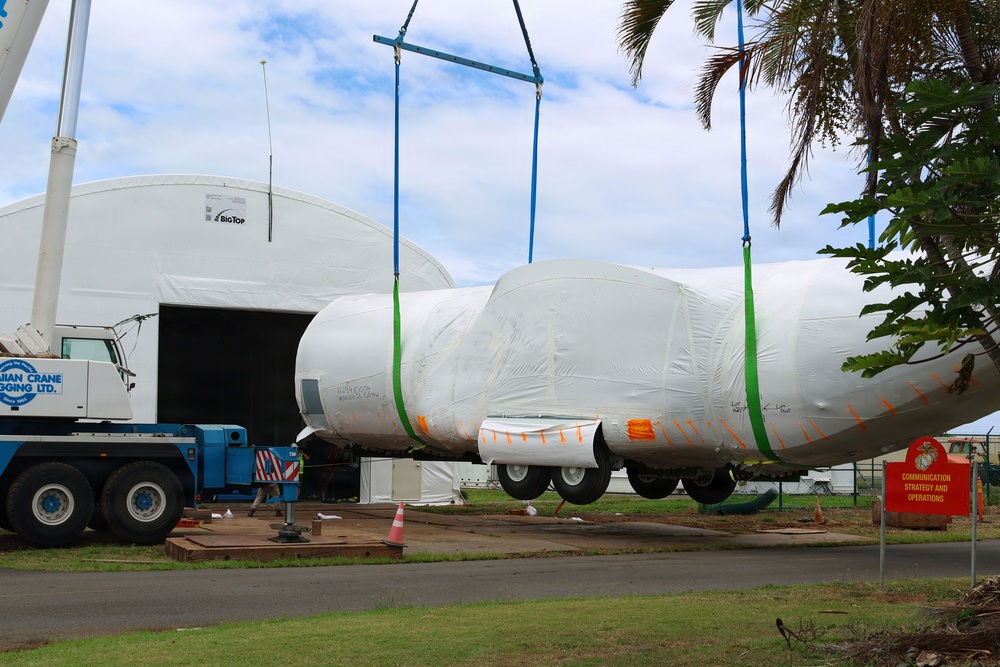 A large aircraft fuselage, wrapped in white protective shrink wrap with orange and green strapping, is being lifted by a crane at what appears to be a military or aviation facility. The aircraft is suspended by blue and green lifting straps attached to a spreader bar connected to a crane. A white temporary structure branded 'Big Top' serves as a hangar in the background, and a red US Marine Corps sign is visible to the right.