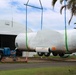 A large aircraft fuselage, wrapped in white protective shrink wrap with orange and green strapping, is being lifted by a crane at what appears to be a military or aviation facility. The aircraft is suspended by blue and green lifting straps attached to a spreader bar connected to a crane. A white temporary structure branded 'Big Top' serves as a hangar in the background, and a red US Marine Corps sign is visible to the right.