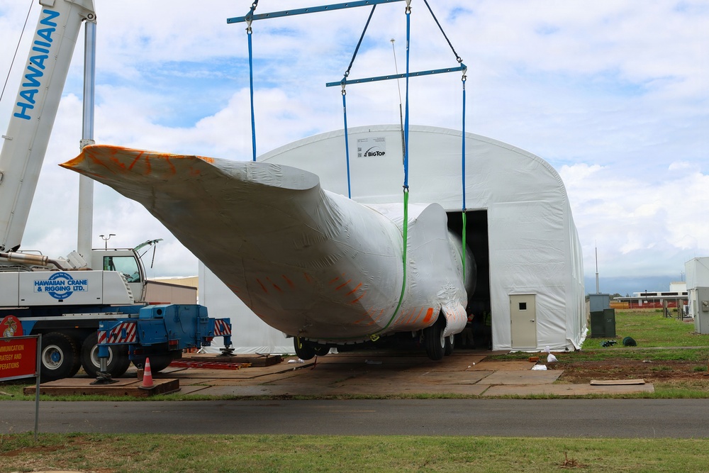 A large wrapped object, likely an aircraft wing or fuselage section, is being lifted by a crane and moved into or out of a temporary fabric structure labeled 'BigTop'. The operation is being conducted by Hawaiian Crane &amp; Rigging Ltd., whose truck and crane boom are visible on the left side of the image. The wrapped component shows orange markings and is secured with blue and green lifting straps attached to a spreader bar.