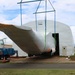 A large wrapped object, likely an aircraft wing or fuselage section, is being lifted by a crane and moved into or out of a temporary fabric structure labeled 'BigTop'. The operation is being conducted by Hawaiian Crane &amp; Rigging Ltd., whose truck and crane boom are visible on the left side of the image. The wrapped component shows orange markings and is secured with blue and green lifting straps attached to a spreader bar.