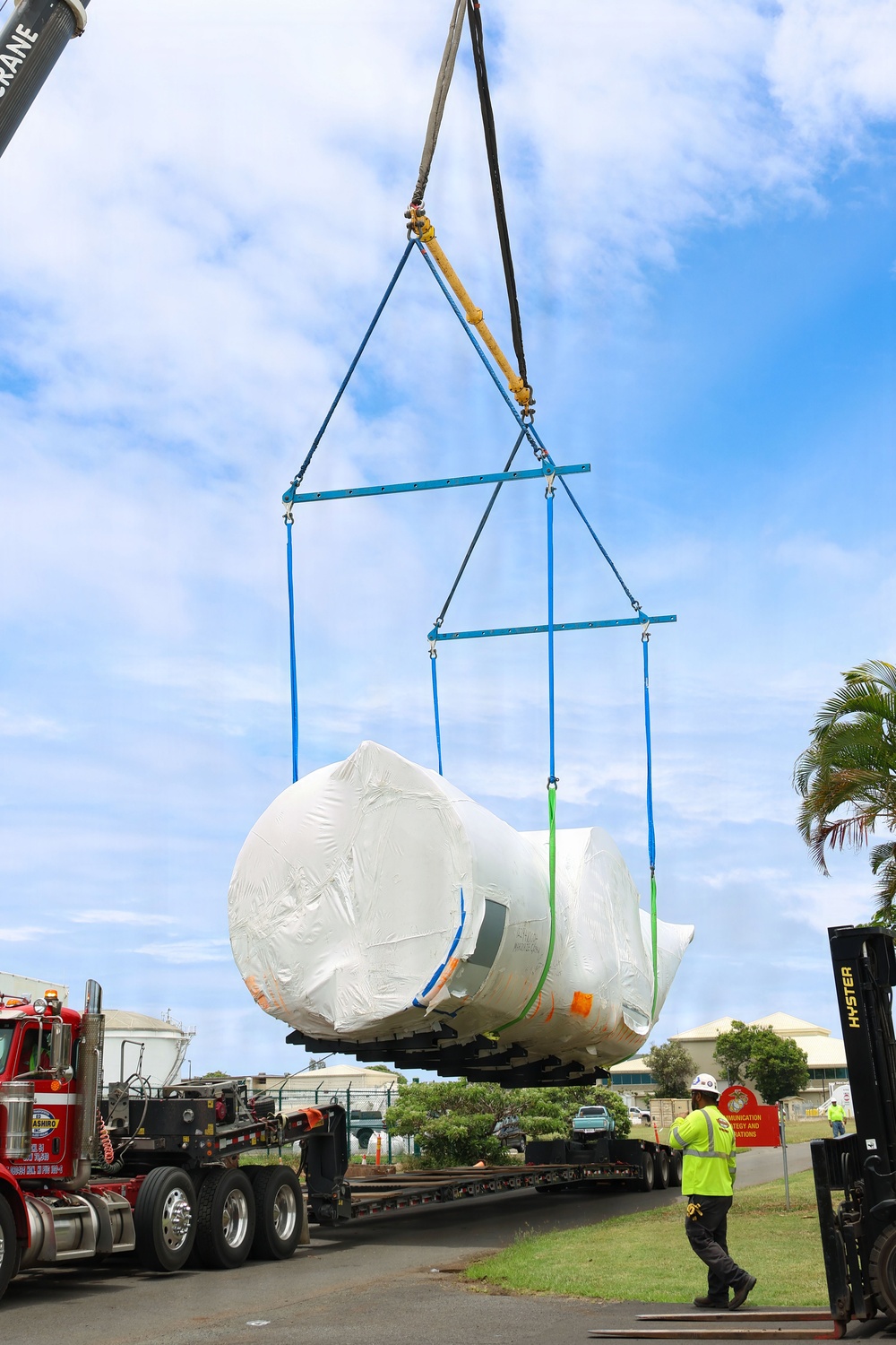 A large crane is lifting a massive shrink-wrapped industrial component — likely a turbine, pressure vessel, or similar heavy equipment — off a flatbed semi-trailer truck. The lift is being managed using a blue spreader bar rigging system with multiple slings. A worker in high-visibility gear and a hard hat observes the operation from ground level, while a Hyster forklift is visible on the right.
