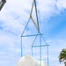 A large crane is lifting a massive shrink-wrapped industrial component — likely a turbine, pressure vessel, or similar heavy equipment — off a flatbed semi-trailer truck. The lift is being managed using a blue spreader bar rigging system with multiple slings. A worker in high-visibility gear and a hard hat observes the operation from ground level, while a Hyster forklift is visible on the right.