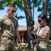 Two military personnel in OCP (Operational Camouflage Pattern) uniforms are working together outdoors, assembling or adjusting what appears to be a military antenna or communications mast mounted on a tripod. Both individuals are smiling, suggesting a positive training or exercise environment. The setting appears to be a military installation or training area with trees and a building visible in the background.