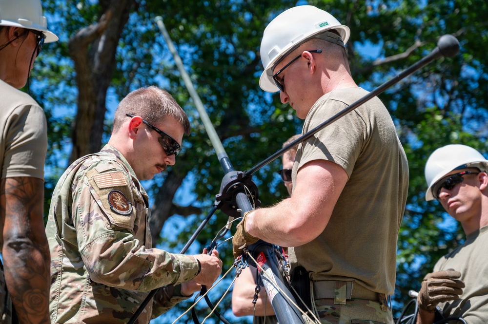 Military personnel in U.S. Air Force uniforms and civilian workers wearing white hard hats are collaborating on assembling or rigging antenna or communications equipment outdoors. The scene appears to be a field training or operational setup involving antenna mast assembly with ropes and rigging hardware. The activity takes place in a sunny, tree-lined outdoor environment.