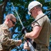 Military personnel in U.S. Air Force uniforms and civilian workers wearing white hard hats are collaborating on assembling or rigging antenna or communications equipment outdoors. The scene appears to be a field training or operational setup involving antenna mast assembly with ropes and rigging hardware. The activity takes place in a sunny, tree-lined outdoor environment.