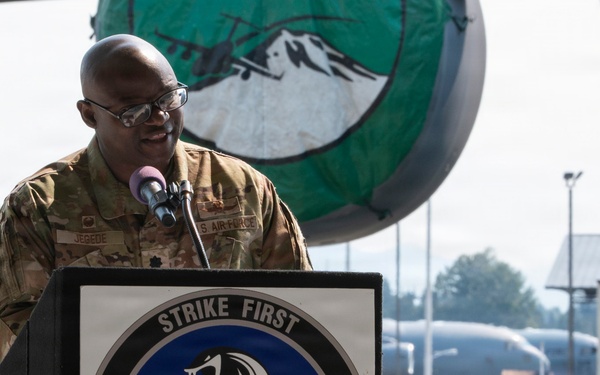 A U.S. Air Force officer in OCP (Operational Camouflage Pattern) uniform is speaking at a podium bearing the insignia of the 627th Communications Squadron with the motto 'Strike First.' The event appears to be an official military ceremony taking place on a flight line, with military aircraft and base infrastructure visible in the background. The podium emblem features a cobra snake with lightning bolts, consistent with the squadron's official patch.