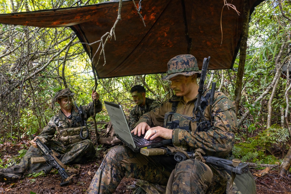 Three military personnel in camouflage uniforms are positioned in a forested area under a makeshift tarp shelter. The foreground soldier is operating a ruggedized laptop computer while armed, suggesting a field command or communications operation. All personnel have face paint applied and are equipped with tactical gear and weapons.