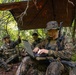Three military personnel in camouflage uniforms are positioned in a forested area under a makeshift tarp shelter. The foreground soldier is operating a ruggedized laptop computer while armed, suggesting a field command or communications operation. All personnel have face paint applied and are equipped with tactical gear and weapons.