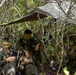 Three military personnel in camouflage uniforms are positioned in a dense jungle or forest environment under a camouflage tarp shelter. They appear to be at a field operations post, with one soldier operating what appears to be a laptop or communications device. The soldiers have green face paint applied, indicating a field exercise or tactical operation.