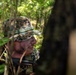 A male individual wearing digital camouflage uniform and matching boonie hat is positioned in dense woodland vegetation, partially concealed by foliage. He appears to be in a surveillance or observation posture, looking to his right. A firearm barrel is partially visible at the lower portion of the frame.