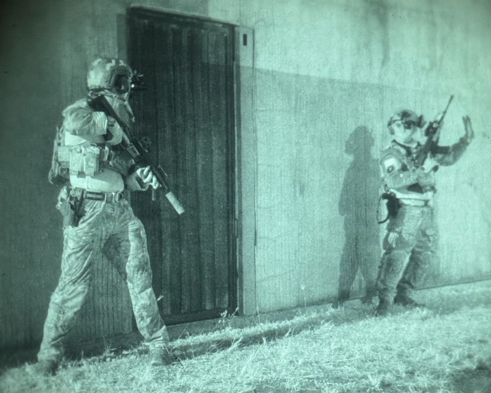 A night-vision captured image showing two armed military or law enforcement personnel conducting a tactical door-breach operation on a building exterior. The left operator is positioned at the door with weapon raised, while the right operator is stacked against the wall providing hand signals. The green-tinted imagery is characteristic of night-vision optics or thermal imaging equipment.