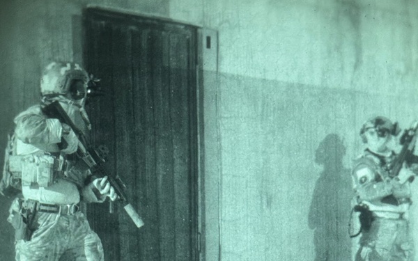 A night-vision captured image showing two armed military or law enforcement personnel conducting a tactical door-breach operation on a building exterior. The left operator is positioned at the door with weapon raised, while the right operator is stacked against the wall providing hand signals. The green-tinted imagery is characteristic of night-vision optics or thermal imaging equipment.
