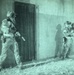 A night-vision captured image showing two armed military or law enforcement personnel conducting a tactical door-breach operation on a building exterior. The left operator is positioned at the door with weapon raised, while the right operator is stacked against the wall providing hand signals. The green-tinted imagery is characteristic of night-vision optics or thermal imaging equipment.