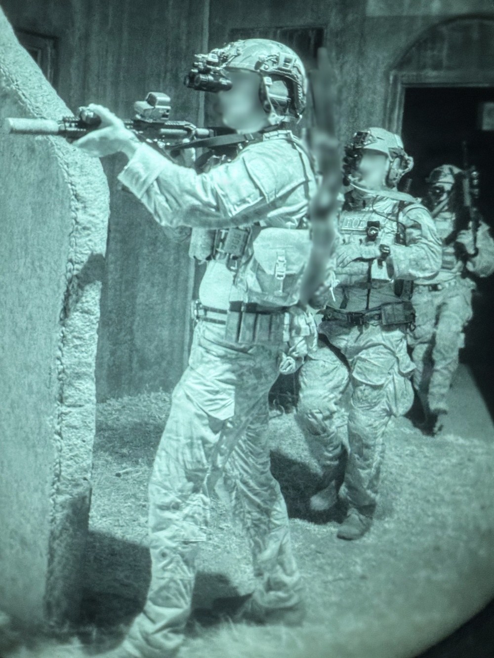A night-vision captured image showing a team of heavily equipped tactical operators conducting what appears to be a building clearance or raid operation. The green-tinted imagery is characteristic of night-vision device (NVD) photography. At least three personnel are visible in a stacked formation near a structure wall, with the lead operator aiming a rifle around a corner.