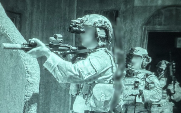 A night-vision captured image showing a team of heavily equipped tactical operators conducting what appears to be a building clearance or raid operation. The green-tinted imagery is characteristic of night-vision device (NVD) photography. At least three personnel are visible in a stacked formation near a structure wall, with the lead operator aiming a rifle around a corner.