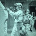 A night-vision captured image showing a team of heavily equipped tactical operators conducting what appears to be a building clearance or raid operation. The green-tinted imagery is characteristic of night-vision device (NVD) photography. At least three personnel are visible in a stacked formation near a structure wall, with the lead operator aiming a rifle around a corner.