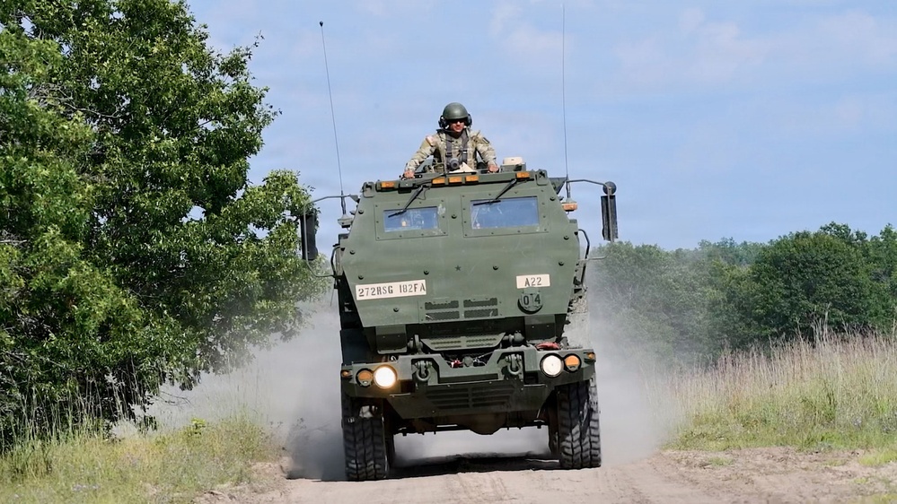 A military wheeled armored vehicle is traveling at speed along a dirt road, kicking up dust. A soldier in combat helmet and gear is visible standing in the open top hatch. The vehicle bears unit markings '272HSG 182FA' and tactical identifier 'A22', consistent with a U.S. Army field exercise.