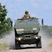 A military wheeled armored vehicle is traveling at speed along a dirt road, kicking up dust. A soldier in combat helmet and gear is visible standing in the open top hatch. The vehicle bears unit markings '272HSG 182FA' and tactical identifier 'A22', consistent with a U.S. Army field exercise.