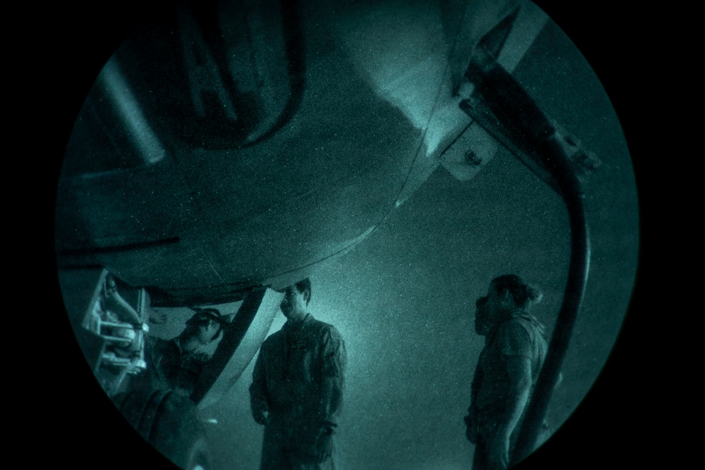 A night-vision or green-tinted circular lens view showing three individuals standing beneath what appears to be a large aircraft or military vehicle. The image has a distinctive fisheye or porthole-style circular framing with heavy vignetting around the edges. The scene appears to be captured at night or in low-light conditions, with a grainy, teal-green monochromatic appearance.