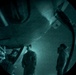 A night-vision or green-tinted circular lens view showing three individuals standing beneath what appears to be a large aircraft or military vehicle. The image has a distinctive fisheye or porthole-style circular framing with heavy vignetting around the edges. The scene appears to be captured at night or in low-light conditions, with a grainy, teal-green monochromatic appearance.