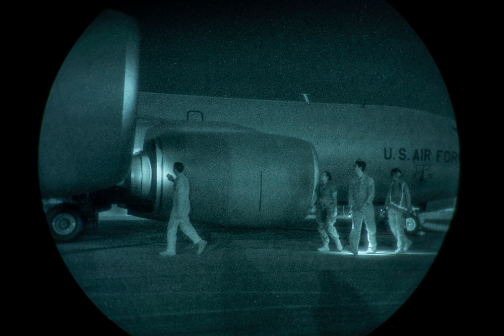 A night-vision captured image showing personnel on a tarmac beside a large U.S. Air Force aircraft. The circular vignette and green-teal tonal range are characteristic of night-vision optics or image intensification equipment. Four individuals in light-colored uniforms or flight suits are visible near the fuselage and engine nacelle of the aircraft.