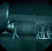 A night-vision captured image showing personnel on a tarmac beside a large U.S. Air Force aircraft. The circular vignette and green-teal tonal range are characteristic of night-vision optics or image intensification equipment. Four individuals in light-colored uniforms or flight suits are visible near the fuselage and engine nacelle of the aircraft.