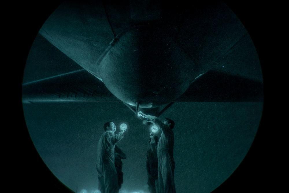 A night-vision or low-light circular-vignette photograph showing two personnel in flight suits conducting an under-aircraft inspection at night. They are using handheld lights to illuminate the underside of what appears to be a large military aircraft. The teal-green color cast and circular frame suggest the image was captured through night-vision optics.