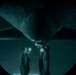 A night-vision or low-light circular-vignette photograph showing two personnel in flight suits conducting an under-aircraft inspection at night. They are using handheld lights to illuminate the underside of what appears to be a large military aircraft. The teal-green color cast and circular frame suggest the image was captured through night-vision optics.