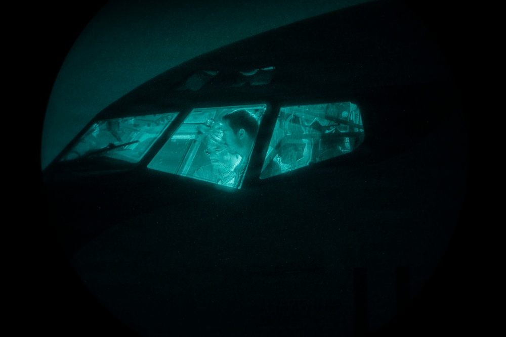 The image appears to be taken underwater or in very low-light conditions, showing the cockpit or cabin windows of what appears to be a submerged aircraft or vessel. Through the multi-pane windows, at least one human figure is visible inside, illuminated by a teal/cyan ambient light. The overall scene is extremely dark with the interior light source creating a dramatic contrast against the surrounding darkness.