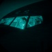The image appears to be taken underwater or in very low-light conditions, showing the cockpit or cabin windows of what appears to be a submerged aircraft or vessel. Through the multi-pane windows, at least one human figure is visible inside, illuminated by a teal/cyan ambient light. The overall scene is extremely dark with the interior light source creating a dramatic contrast against the surrounding darkness.