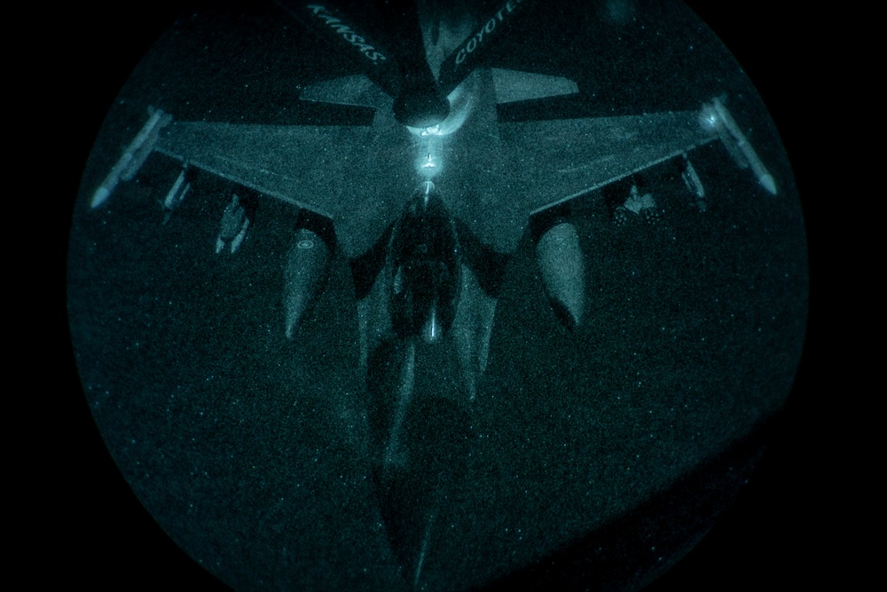 A night-vision or low-light aerial photograph showing a military aircraft viewed from above through what appears to be a boom operator's window of a tanker aircraft during an aerial refueling operation. The circular vignette framing is characteristic of a refueling boom pod observation window. Text markings including 'KANSAS' and 'COYOTES' are faintly visible on the tanker aircraft's tail section in the upper portion of the frame.