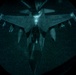 A night-vision or low-light aerial photograph showing a military aircraft viewed from above through what appears to be a boom operator's window of a tanker aircraft during an aerial refueling operation. The circular vignette framing is characteristic of a refueling boom pod observation window. Text markings including 'KANSAS' and 'COYOTES' are faintly visible on the tanker aircraft's tail section in the upper portion of the frame.
